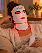 Woman relaxing on a sofa wearing the illuminated Face + Neck Mask with its controller visible